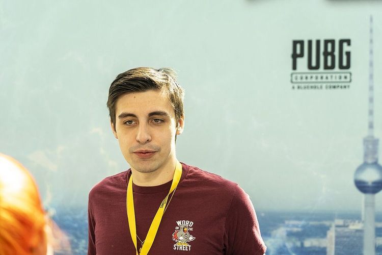 A young man with short brown hair in a maroon T-shirt and yellow lanyard stands in front of a PUBG event backdrop.