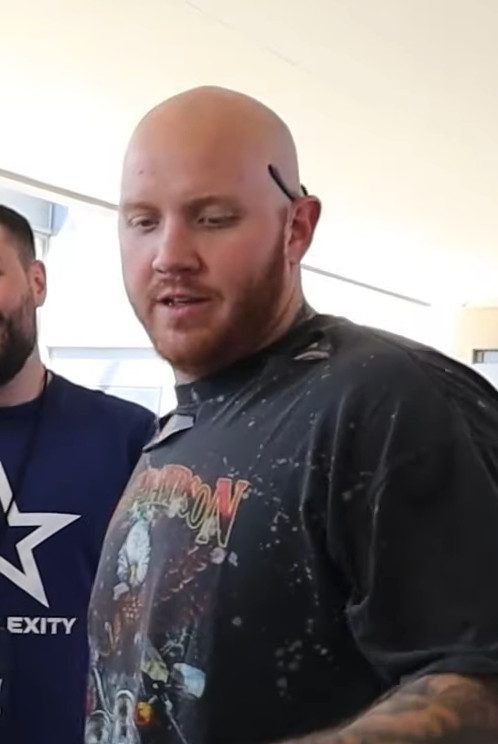 A bald man with a red beard in a black graphic T-shirt speaks to someone out of frame.