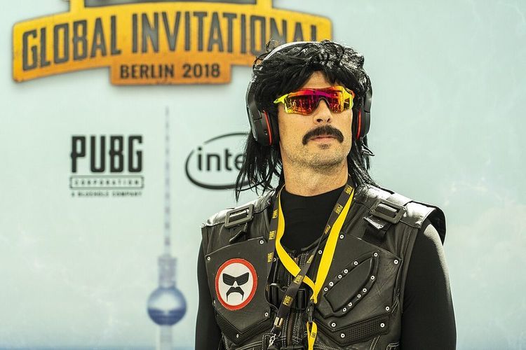 A man in a black mullet wig, mirrored sunglasses, headphones and a black vest stands in front of a PUBG Global Invitational Berlin 2018 banner.