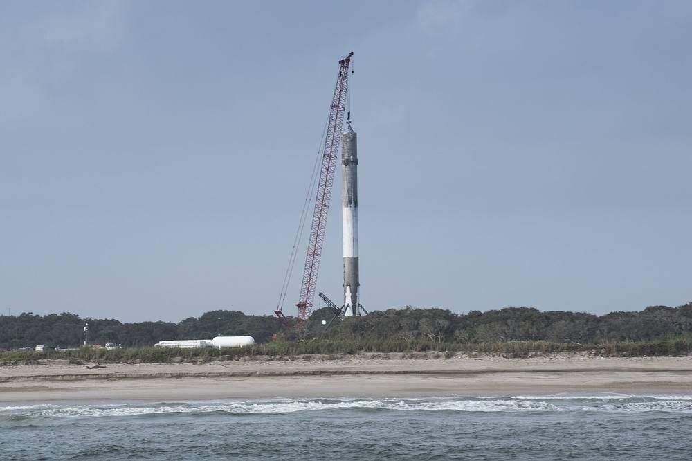 A tall white rocket booster with landing legs stands on a concrete pad beside the sea at Cape Canaveral under a pale sky.
