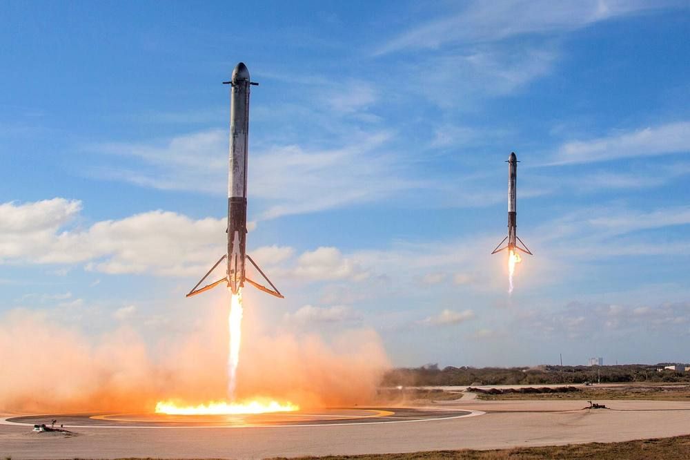 Two white booster stages descend side by side on flames toward adjacent landing pads on the coast.