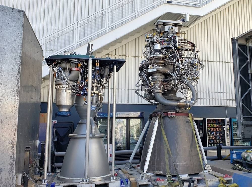 Two rocket engines with large bells stand side by side on stands in a factory hall, the right one with a more complex tangle of plumbing.