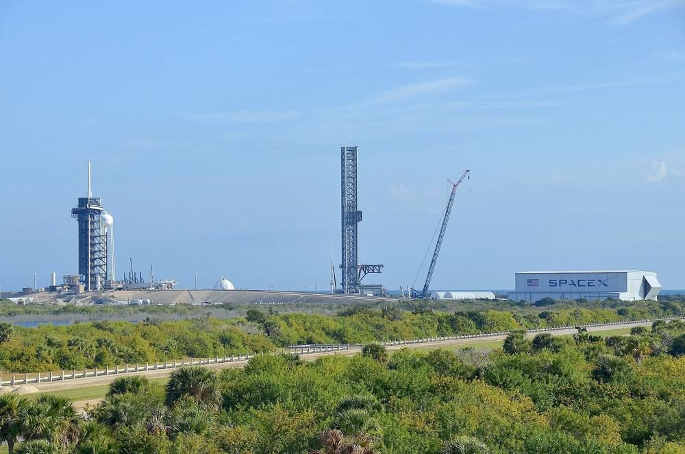 A flat green Florida landscape under a blue sky with a launch tower and a tall steel Starship tower in the middle distance.