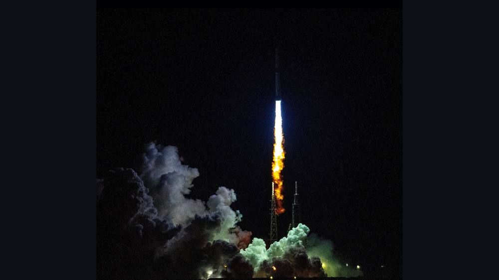 A large rocket climbs from a launch pad on a column of fire against a dark sky with a cloud of exhaust below.