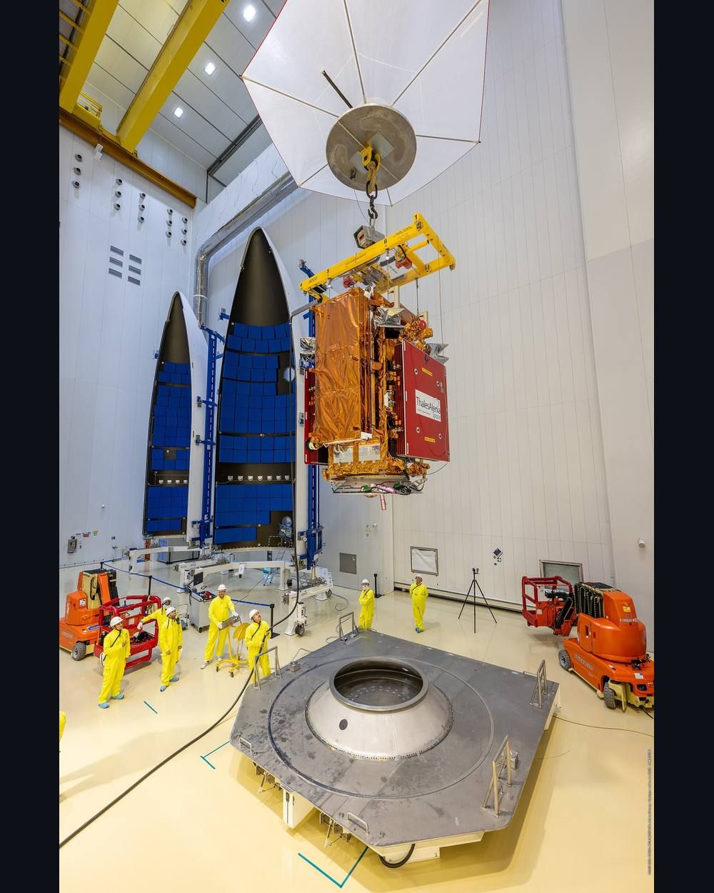 Technicians in yellow suits work around a satellite hanging from a crane above the adapter of a launch vehicle in a bright clean room.