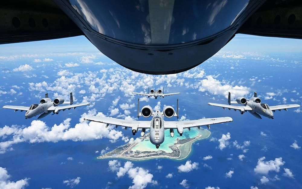 Four A-10 jets in formation above scattered cloud and a small ringed island, with the underside of a refueling aircraft at the top of the frame.
