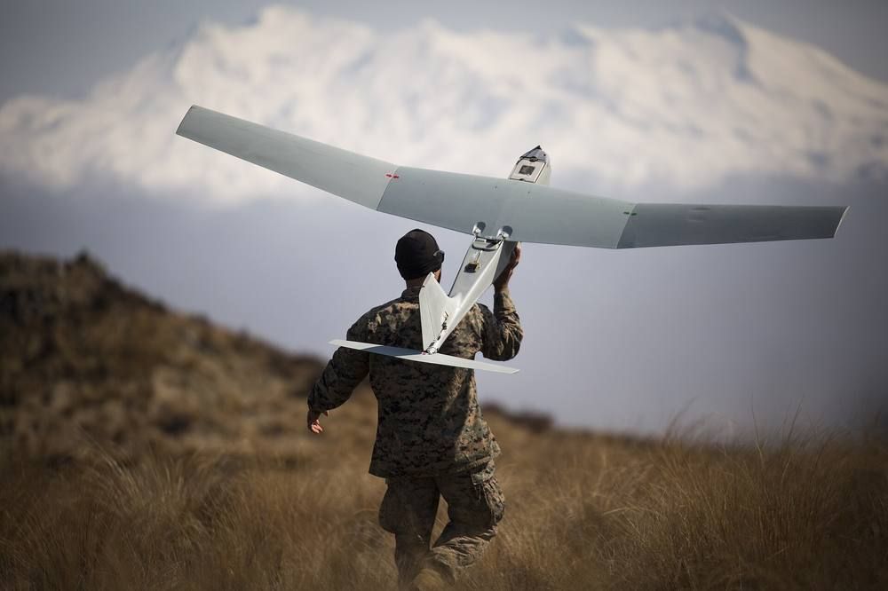A person in camouflage uniform, seen from behind on a grassy hillside, carrying a small white long-winged drone at shoulder height before launching it.